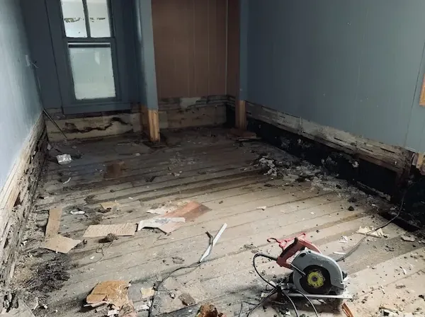 A room with the baseboard and lower wall cut away after a pipe leak, a saw resting on the bare floor
